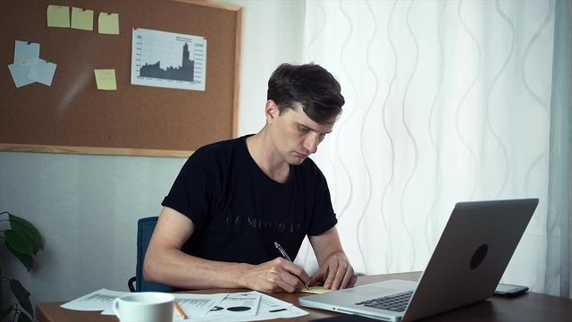 Young man writing ideas on adhesive notes and attaching it to board at office. Freelancer working on laptop computer in the home office.