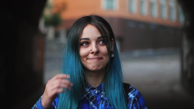Street punk or hipster girl with blue dyed hair. Woman with piercing in nose, violet lenses, ears tunnels and unusual hairstyle stands in city. Slow motion.