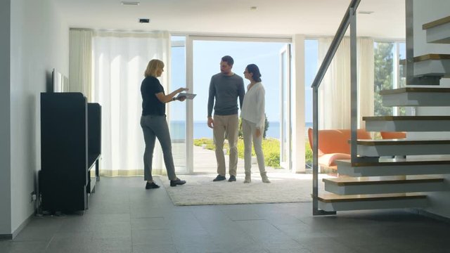 Professional Real Estate Agent Shows Stylish Modern House to a Beautiful Young Couple Who are in the Market for Purchasing/ Renting New Home. House Has Floor to Ceiling Windows and Seaside View.