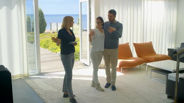Professional Real Estate Agent Shows Stylish Modern House to a Beautiful Young Couple Who are in the Market for Purchasing/ Renting New Home. House Has Floor to Ceiling Windows and Seaside View. 