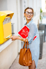 Young woman putting letter to the old yellow mailbox standing with bag and magazine outdoors on the...