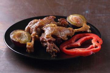 Fried meat with plums and red bell pepper on a black plate with a wooden fork on a rusty background