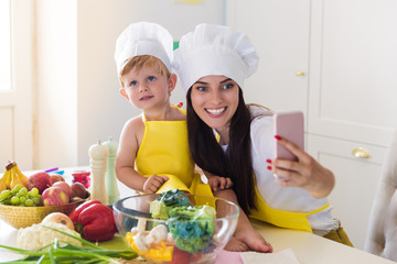 The young smiling cook mother sitting with her cute little son in the kitchen near vegetables and doing selfie-photo on the phone