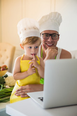 Portrait of cute little boy who sitting in cook form on a table in the kitchen near smiling father in cook form and they looking at laptop