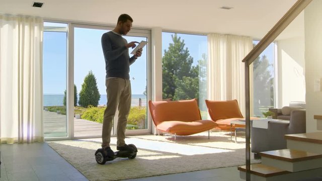 Tech Industry Businessman Working From Home Uses Tablet Computer and Rides Around the House on His Gyro Scooter. His Modern House has Seaside View.