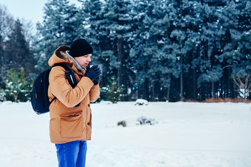 a young man in a red jacket with a backpack standing on the snow and holding a thermos and a mug of tea. Winter forest.
