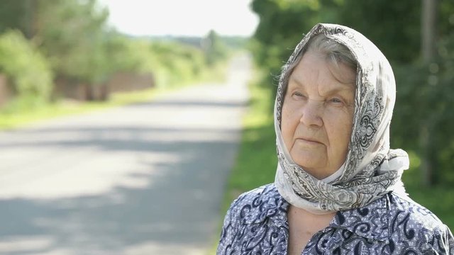 Thoughtful look of a serious mature old woman with gray hair dressed in a handkerchief on the background of road in summer. Slow Motion
