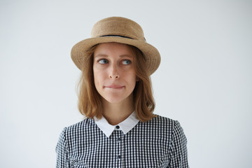 Studio shot of beautiful hesitated young female wearing stylish round hat and dress looking...