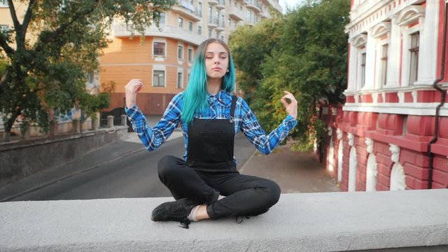 Calm street punk or hipster girl with blue dyed hair. Woman with piercing in nose, unusual hairstyle meditating on European empty street. Yoga concept. Slow motion.