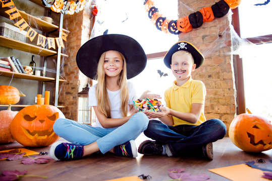 Sweets for us! Treat or trick! Very cheerful excited small kids in carnival head wear, with colorful treats, carved pumpkins, fall leaves, orange papers near them on the brown wooden floor