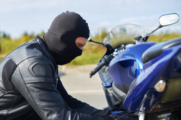 Stylish young Caucasian male biker wearing black leather jacket and mask checking his blue...