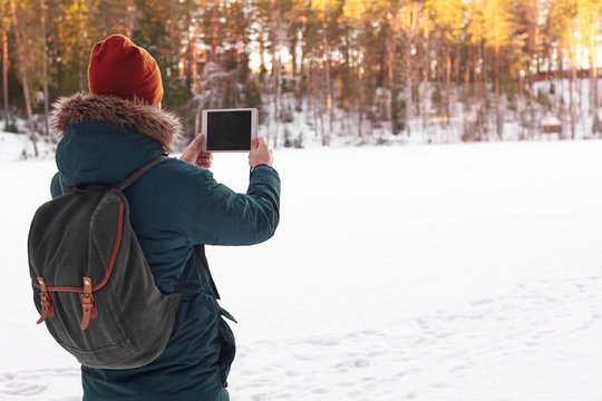 Back view of unrecognizable male in hat and cout with fur hood carrying stylish backpack taking picture of amazing winter landscape using copyspace mobile phone while walking alone on sunny day