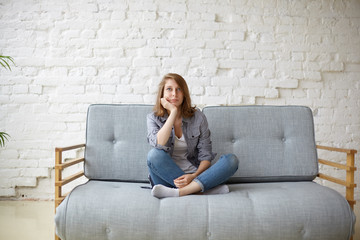 Portrait of beautiful pensive young Caucasian female keeping legs crossed while sitting on couch...