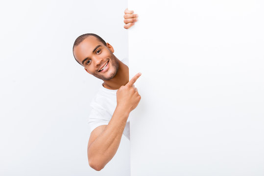 Happy Amazed Young Mulatto Guy Is Popping Out From White Blank Banner, Pointing At It With Finger, Wearing White T Shirt, On White Background