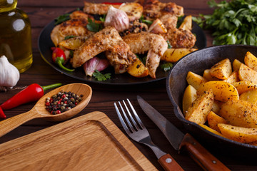 Grilled chicken wings, baked potatoes, herbs and spices on kitchen table. Hot fresh appetizing countryside food. Cooking concept, meat dishes, homemade cuisine