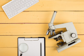 Education and science concept - microscope, magnifying glass, blank clipboard, computer keyboard on the yellow desk