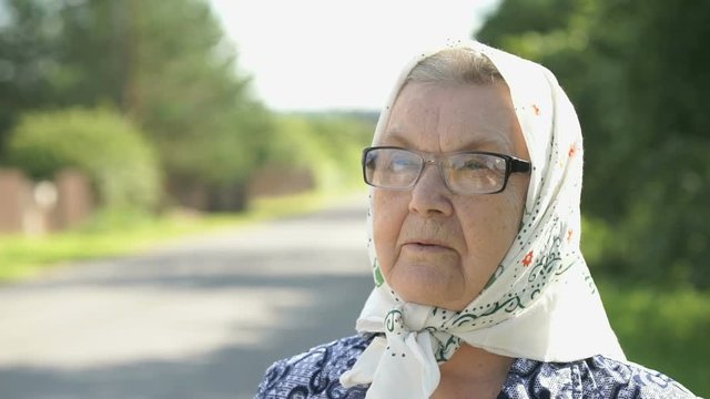 Portrait of serious mature old woman with gray hair in glasses dressed in a white handkerchief on the background of road in summer. Close-up