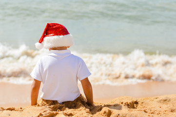 Little child in Santa Claus hat on sundy beach outdoors background. Back view picture