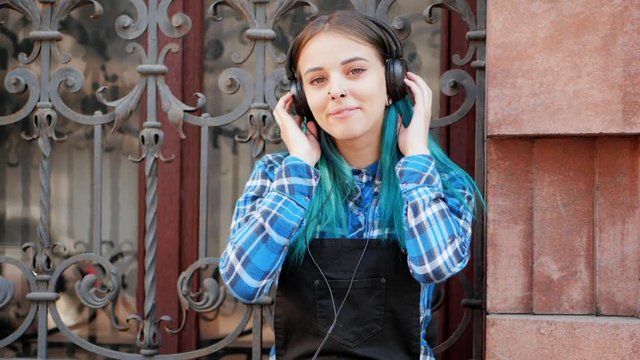 Street punk or hipster girl listening music with brown headphones.Teen girl with blue dyed hair,piercing in nose,violet lenses,unusual hairstyle sitting near historic building.Slow motion.