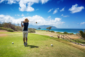 Golfer with straw hat teeing off with driver from ocean side par 4 hole - obrazy, fototapety, plakaty