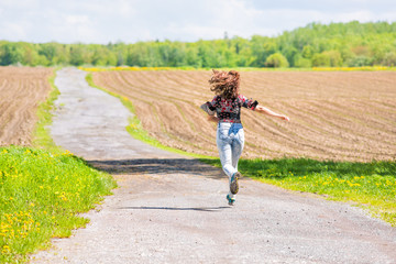 Obraz premium Back of young woman running, jumping in air and smiling on dirt countryside road by brown plowed fields with furrows in summer in Ile D'Orleans, Quebec, Canada