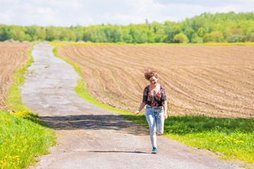 Young woman running, jumping in air and smiling on countryside dirt road by brown plowed fields with furrows in summer in Ile D'Orleans, Quebec, Canada