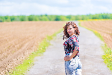 Naklejka na ściany i meble Young woman standing posing in jeans, turned back on countryside dirt road by brown plowed field with furrows in summer in Ile D'Orleans, Quebec, Canada with forest in background