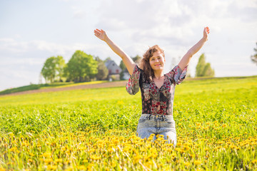 Naklejka na ściany i meble Young happy smiling woman sitting in yellow dandelion flower farm field during sunset by countryside rural house in Ile D'Orleans, Quebec, Canada raising hands