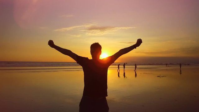 SLOW MOTION, CLOSE UP, LENS FLARE: Excited young man cheering with arms raised on dreamy sandy beach at magical orange sunset. Happy guy lifting hands in the air with excitement. People on holidays