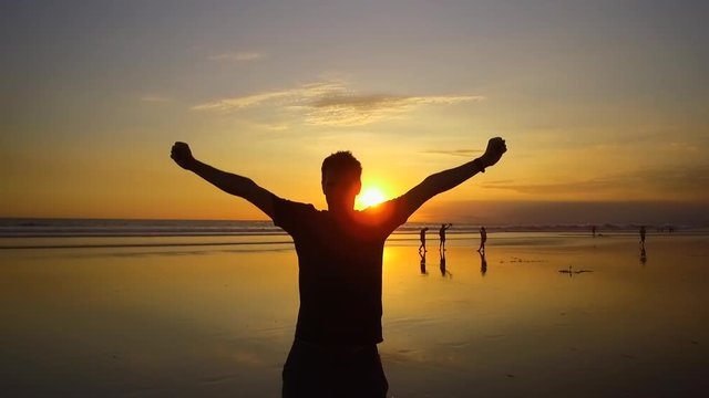 SLOW MOTION CLOSE UP SILHOUETTE Excited young man cheering with arms raised. People on dreamy sandy beach at magical orange sunset. Happy guy on seashore outstretching hands with excitement at sunrise