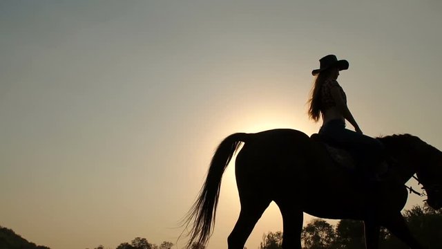 Close-up silhouette of a young girl in a cowboy hat riding a horse on sunset sky background. Slow motion.