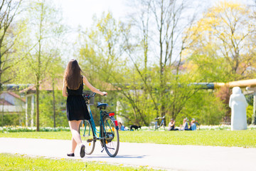 Back view of girl on bicycle wearing on black short dress. Young Woman riding along road on green spring  outdoor Park. Sporty young girl riding a bicycle on a sunny morning, view from the back
