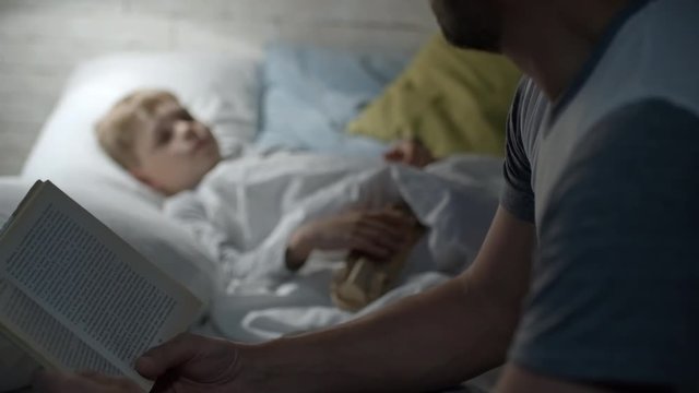 PAN of unrecognizable father sitting in cozy bedroom and reading book to his little son with wooden toy lying in bed and listening