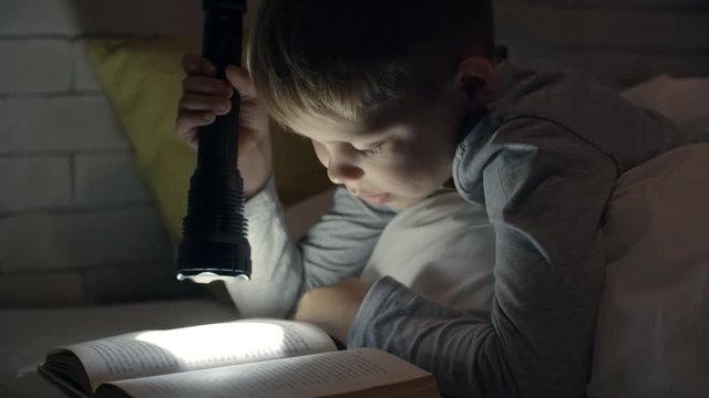 Cute little boy with flashlight lying in bed and reading book in darkness