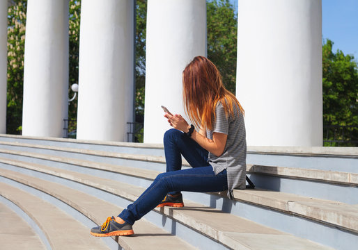 A girl with a phone in her hand writes a message, sitting on the steps.