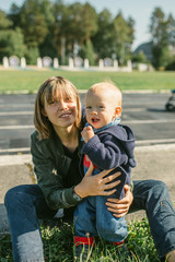 Happy young mother with her son sit on the grass, smile and hug.Outdoors healthy child activity, active lifestyle and having fun on family summer vacation with son at the playground.