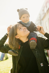 Happy young mother with her son on her shoulder, enjoying the sunshine, they smile and laugh.Outdoors healthy child activity, active lifestyle and having fun on family summer vacation with son at the 