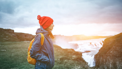 Girl in warm clothing, in red knitted hat and small orange backpack stands on the cliff on background of Gullfoss waterfall in Iceland. Travel to the Iceland