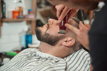 Fototapeta premium Close up shot of a young bearded man getting his beard shaped and trimmed at the barbershop. 