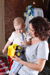 Young mother holds little cute son and toy truck in room, boy looks at toy
