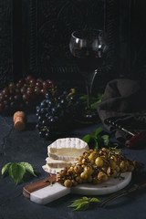 Serving board with sliced camembert cheese and baked bunch of green grapes served with glass of red wine, corkscrew, green leaves over black table. Dark rustic still life