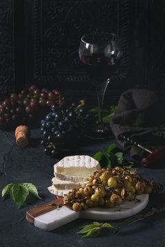 Serving board with sliced camembert cheese and baked bunch of green grapes served with glass of red wine, corkscrew, green leaves over black table. Dark rustic still life