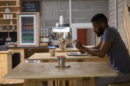 Young man with laptop reading paper while sitting in cafe