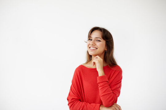 Positive human facial expressions and emotions. Isolated shot of attractive European student girl in red jamper looking and smiling broadly at camera during nice conversation with someone