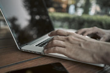 A young man is typing something on a laptop. Sitting in a park at a wooden table