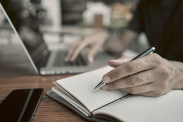 Close-up. A young man writes in a notebook. On the background is a laptop on a wooden table