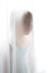 A nice little blond girl stands by the window behind a transparent curtain