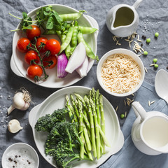 Ingredients for lunch - orzo pasta, asparagus, green peas, broccoli, cherry tomatoes, olive oil, cream and spices. On a gray background, top view