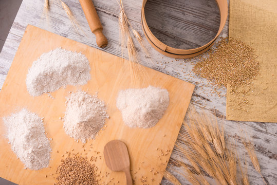 Farine integrali diverse sull&rsquo;asse di legno con chicchi e spighe, setaccio e mattarello