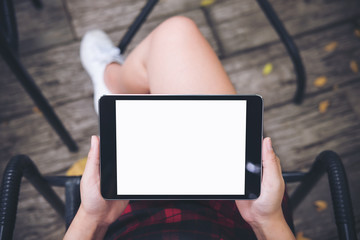 Mockup top view image of woman's hand holding black tablet pc with blank white screen on thigh with wooden floor background in vintage cafe